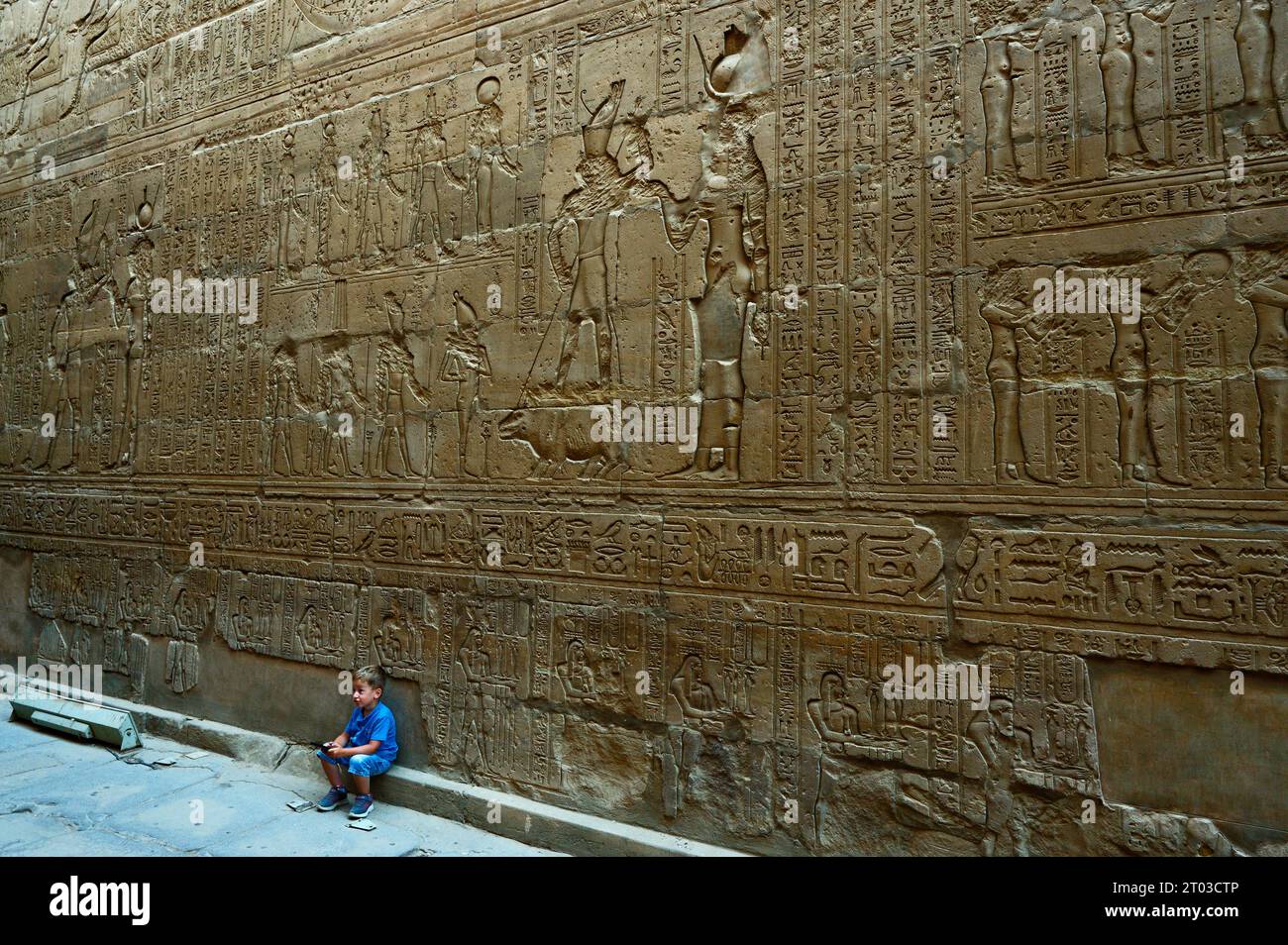 Affascinato dalla meraviglia moderna: Il ragazzo si è arenato nel suo cellulare sullo sfondo di una straordinaria parete geroglifica nel tempio di Edfu. Foto Stock