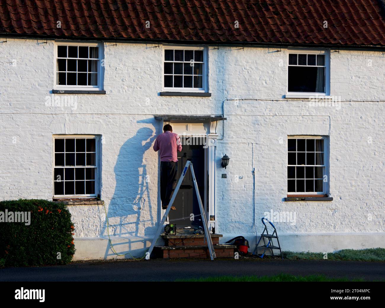 Uomo in piedi sulla scala per riparare la porta della sua casa, nel villaggio di Bishop Wilton, East Yorkshire, Inghilterra, Regno Unito Foto Stock
