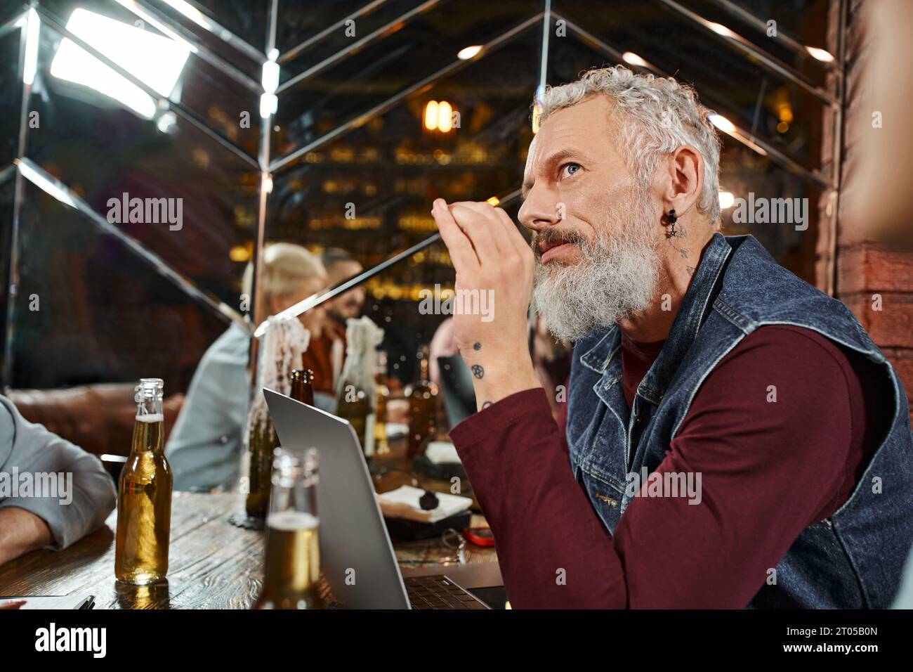 un uomo con la barba premurosa che guarda lontano vicino a un notebook e a colleghi che riflettono allo specchio in un pub Foto Stock