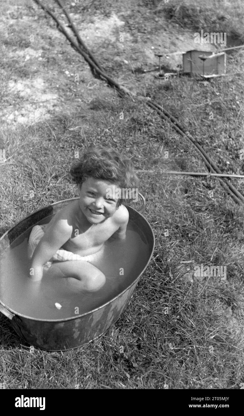 Anni '1950, storico, guardando la telecamera, una dolce bambina seduta in acqua a fare un bagno all'aperto in una vasca di lavaggio in metallo galvanizzato, Inghilterra, Regno Unito. Foto Stock