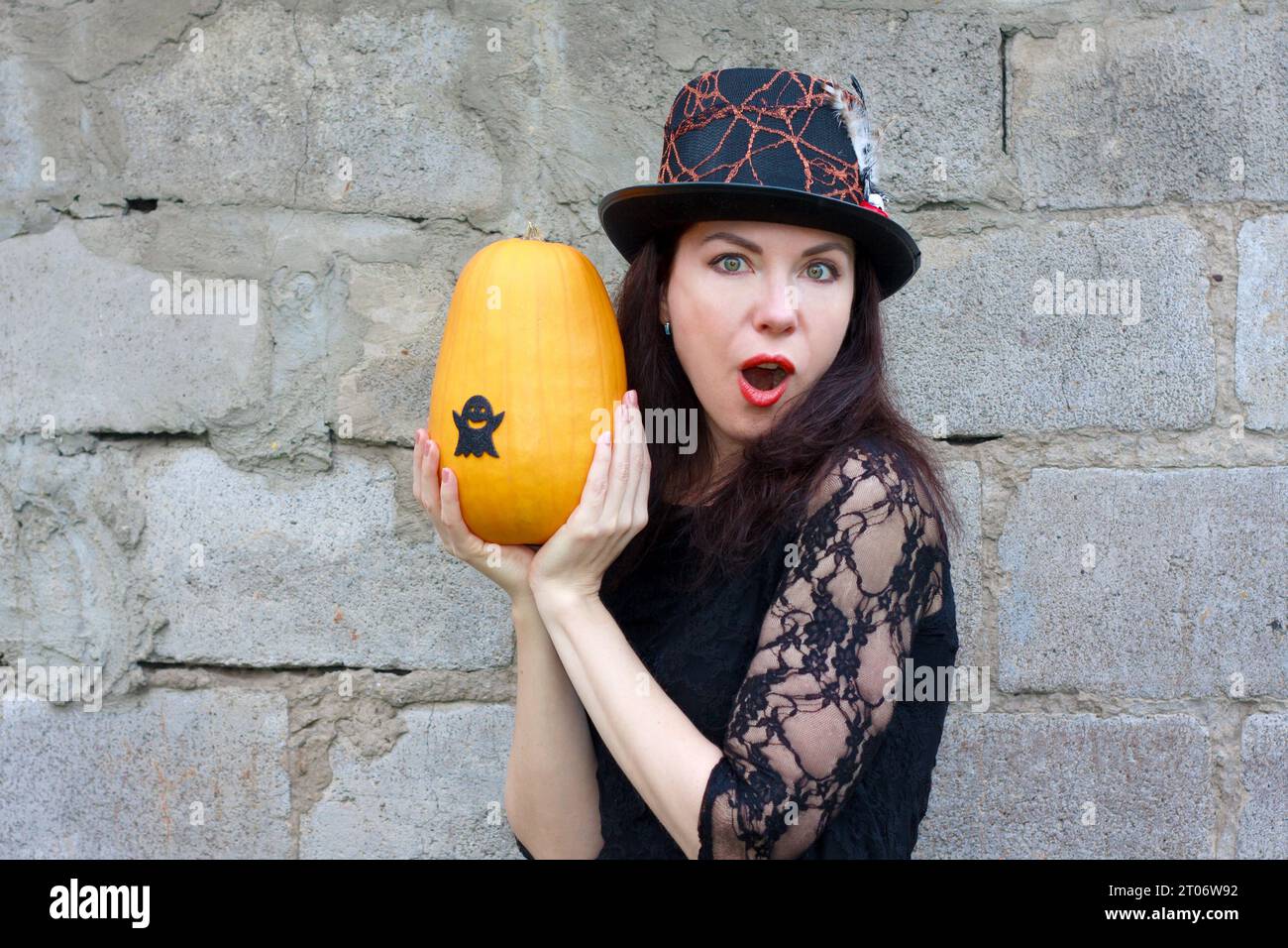 Ritratto ravvicinato di una giovane donna in costume di halloween che esprime sorpresa. donna dai capelli castani con abito nero guipure e cappello che tiene la zucca nel suo han Foto Stock