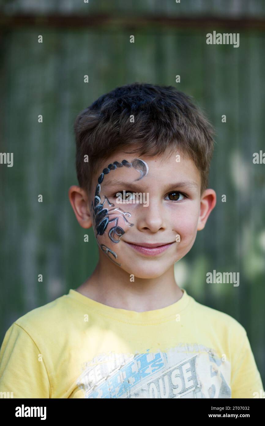 Ritratto di bambino prescolare con scorpione dipinto sul viso. Pittura facciale festiva. Felice bambino dagli occhi castani all'aperto su sfondo verde. Estate. corpo Foto Stock