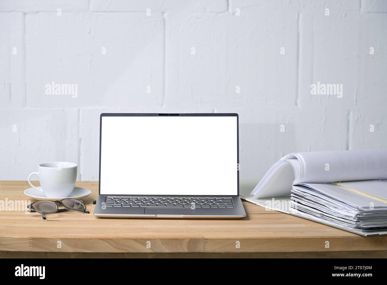 Modello di un computer portatile con schermo bianco vuoto, cartella e tazza da caffè su una scrivania di legno di fronte a una parete dipinta di bianco, ideale per ufficio e busi Foto Stock