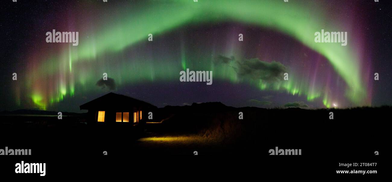 La luminosa e colorata aurora boreale, l'aurora boreale, balla attraverso un cielo aperto con una cabina in primo piano e luci accese nelle finestre Foto Stock