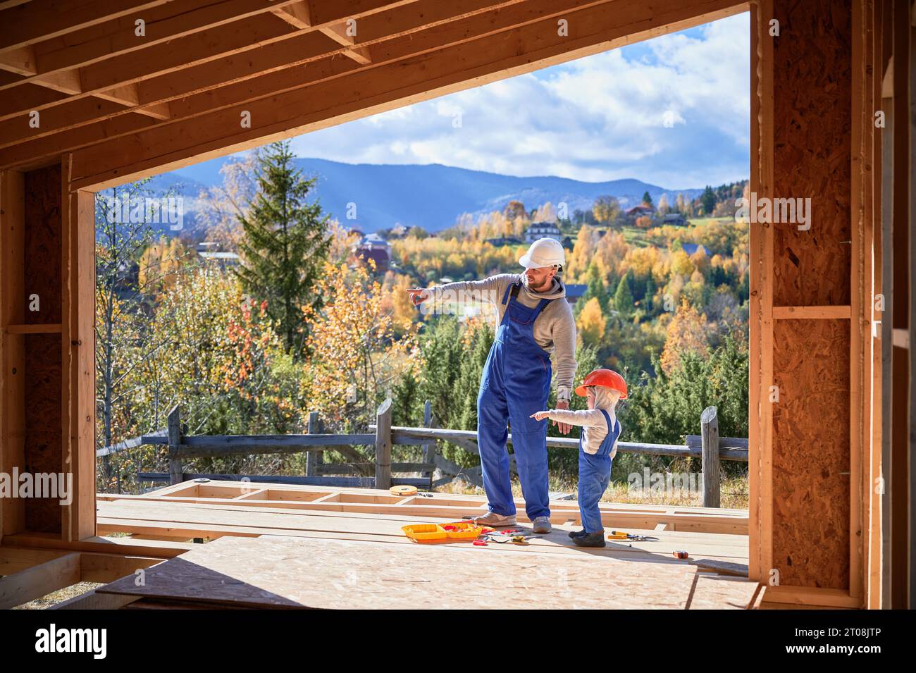 Padre con figlio piccolo costruendo casa con struttura in legno. Costruttore maschile con bambino che punta sulla parete in cantiere, indossando casco e tute blu nelle giornate di sole. Falegnameria e concetto di famiglia. Foto Stock