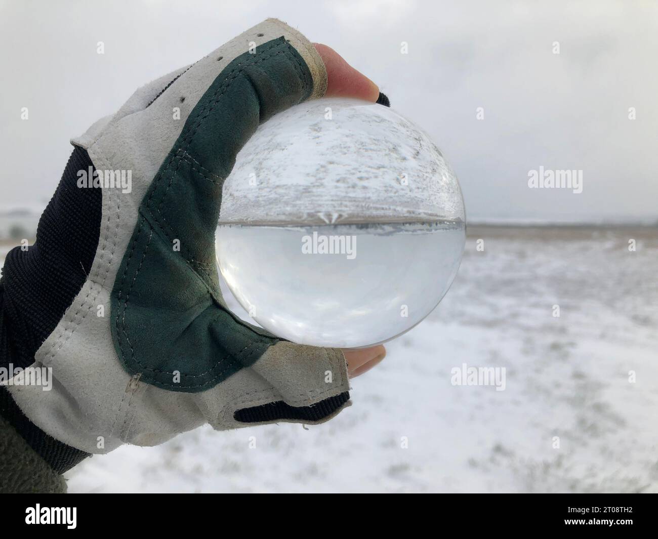 Sfera di cristallo tenuta in una mano con guanti, guardando il paesaggio con neve, orizzonte piatto e cielo nuvoloso in inverno. Fotografia creativa a rifrazione. Foto Stock