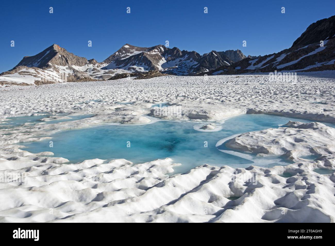 Lago ghiacciato nell'alta Sierra che inizia a sciogliersi a luglio con acqua blu nella neve e montagne all'orizzonte Foto Stock