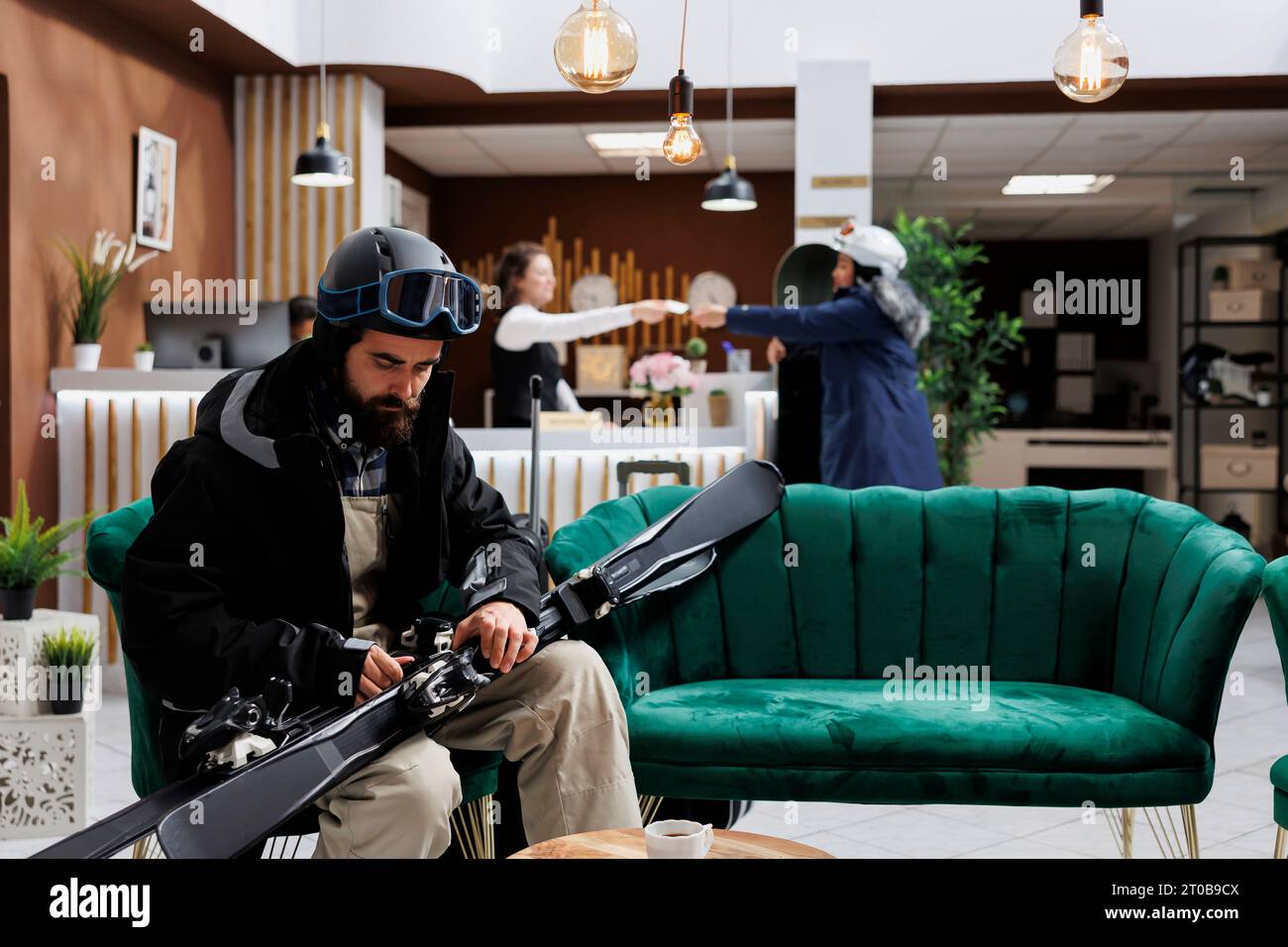 Il turista vestito da giacca invernale controlla l'attrezzatura da sci mentre si siede nella reception dell'hotel. L'uomo con gli sci e con gli occhiali da sci suggerisce di essere pronto per le avventure innevate durante le vacanze invernali. Foto Stock