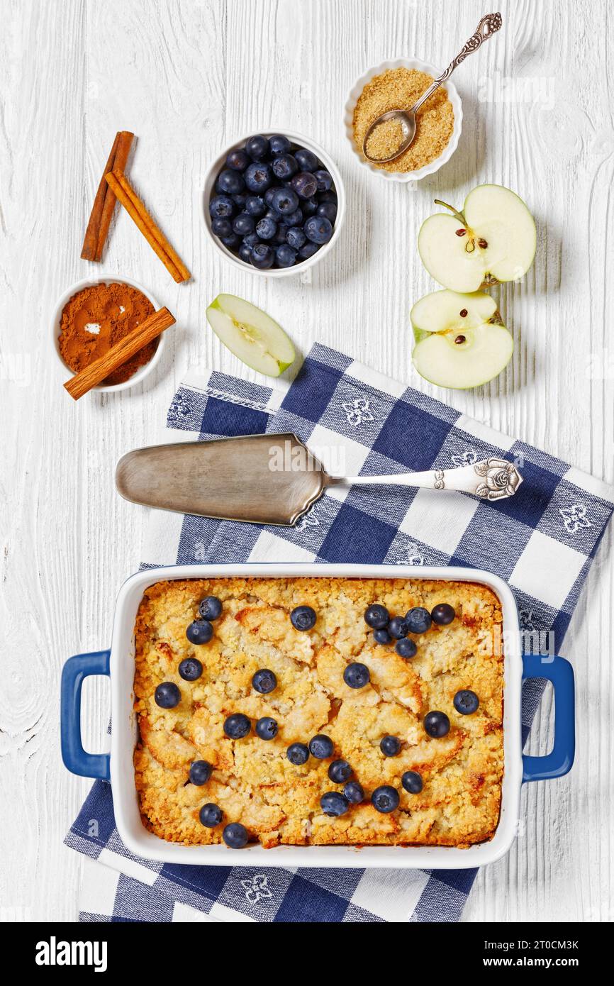 Crumble di mele fatto in casa con mele, cannella, spezie, crumble dolce e burroso e mirtilli freschi cosparsi sopra la piastra da forno su legno bianco Foto Stock