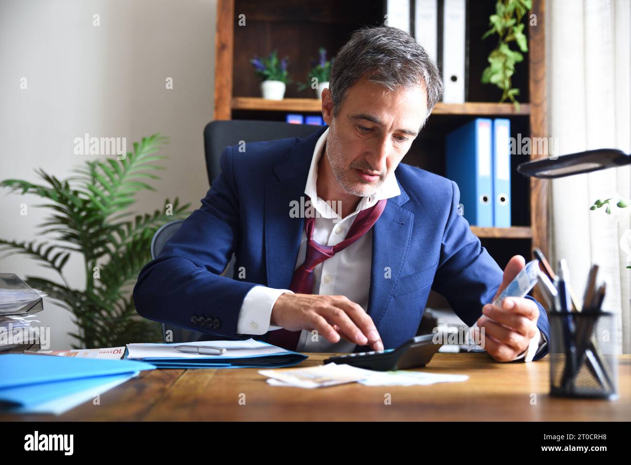 Stressato uomo d'affari che esegue calcoli finanziari su una calcolatrice su un tavolo da ufficio in legno pieno di documenti in un ufficio con scaffali sul retro Foto Stock