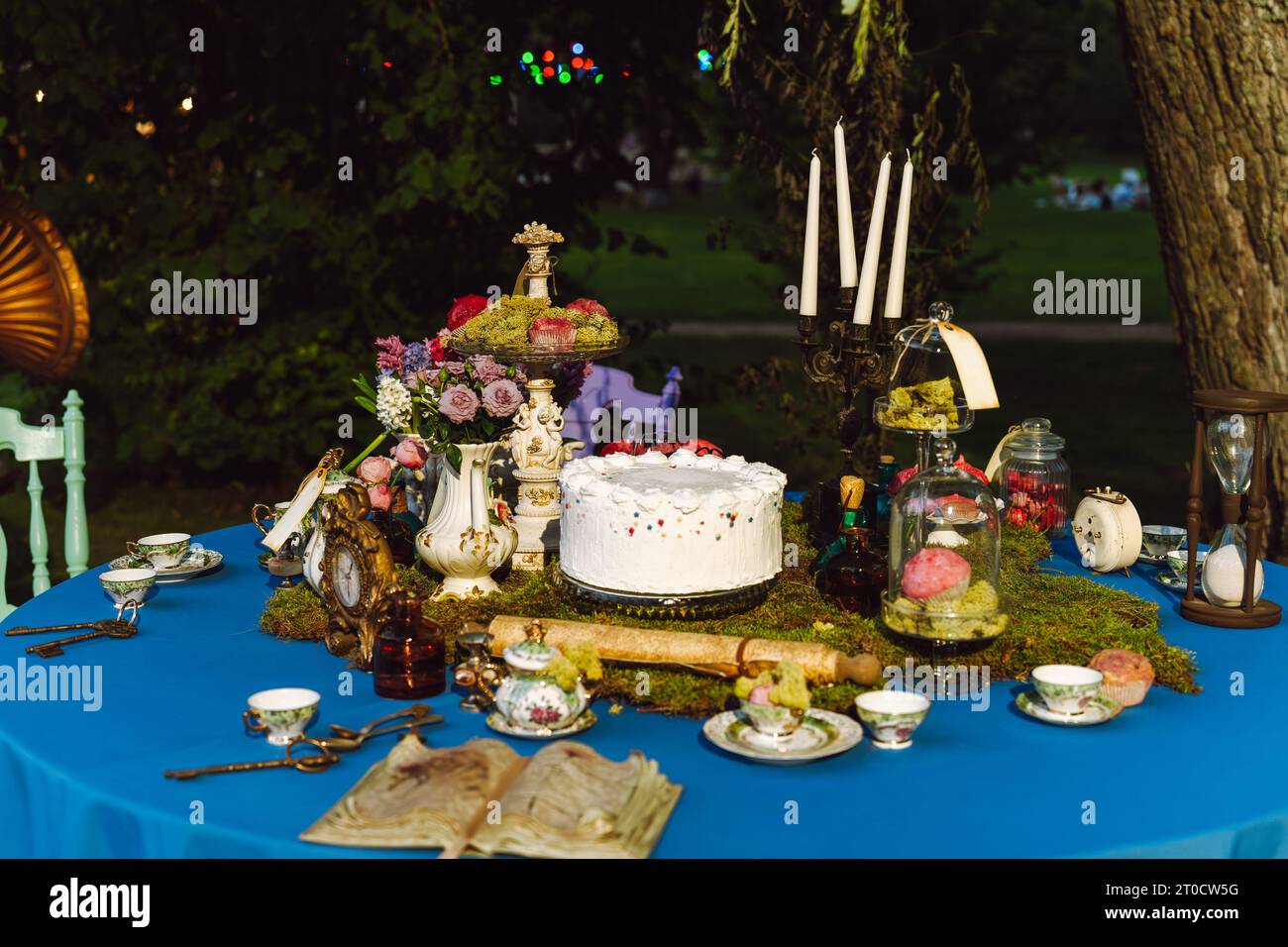 Il tavolo da pranzo e' decorato nello stile di Alice nel Paese delle meraviglie. Un vecchio libro aperto, una torta, ciotole da tè, candele in un candelabro, una clessidra su un Foto Stock