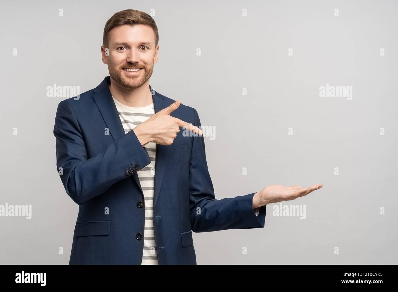 Un uomo d'affari sorridente e barbuto che punta il dito indice a portata di mano sullo spazio di copia su sfondo grigio. Foto Stock