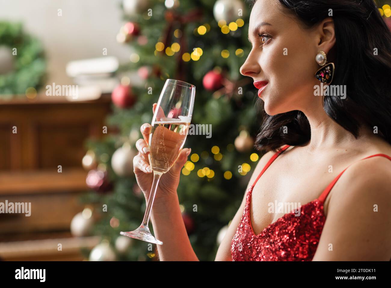 Donna elegante in abito rosso con bicchiere di champagne vicino all'albero di Natale decorato, vita ricca Foto Stock