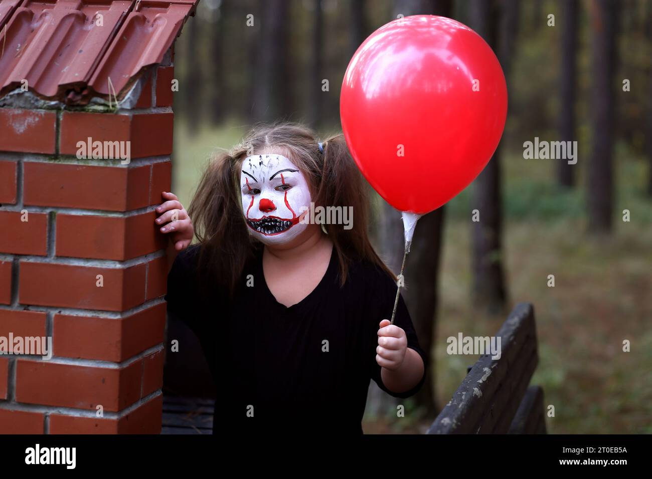 Ragazza seria con un outfit spettrale stilizzata come Pennywise che guarda la fotocamera mentre si trova in un cortile abbandonato con un garage sullo sfondo durante Hal Foto Stock