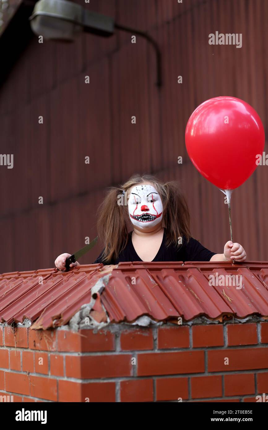 Ragazza seria con un outfit spettrale stilizzata come Pennywise che guarda la fotocamera mentre si trova in un cortile abbandonato con un garage sullo sfondo durante Hal Foto Stock