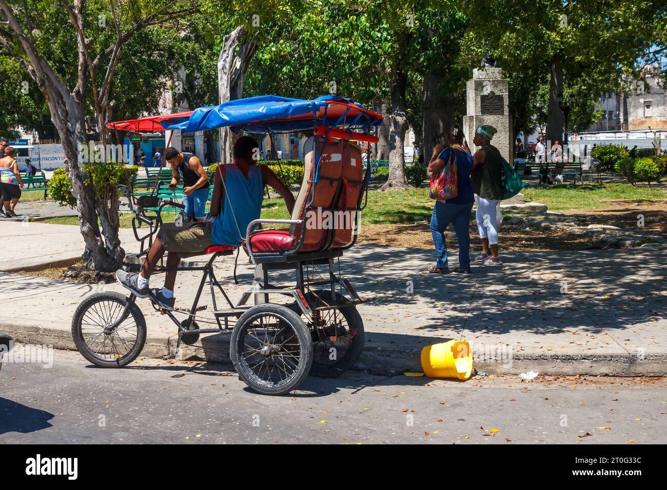 Un autista di pedicab o bicitaxi attende i passeggeri nel quartiere Parque Central. Un secchio di plastica rotto viene gettato nel marciapiede. Foto Stock