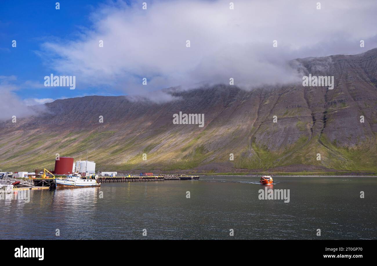 Barca arancione careggiata da turisti che si avvicinano al molo di Isafjordur, in Islanda, con acqua in primo piano e un cielo spettacolare Foto Stock