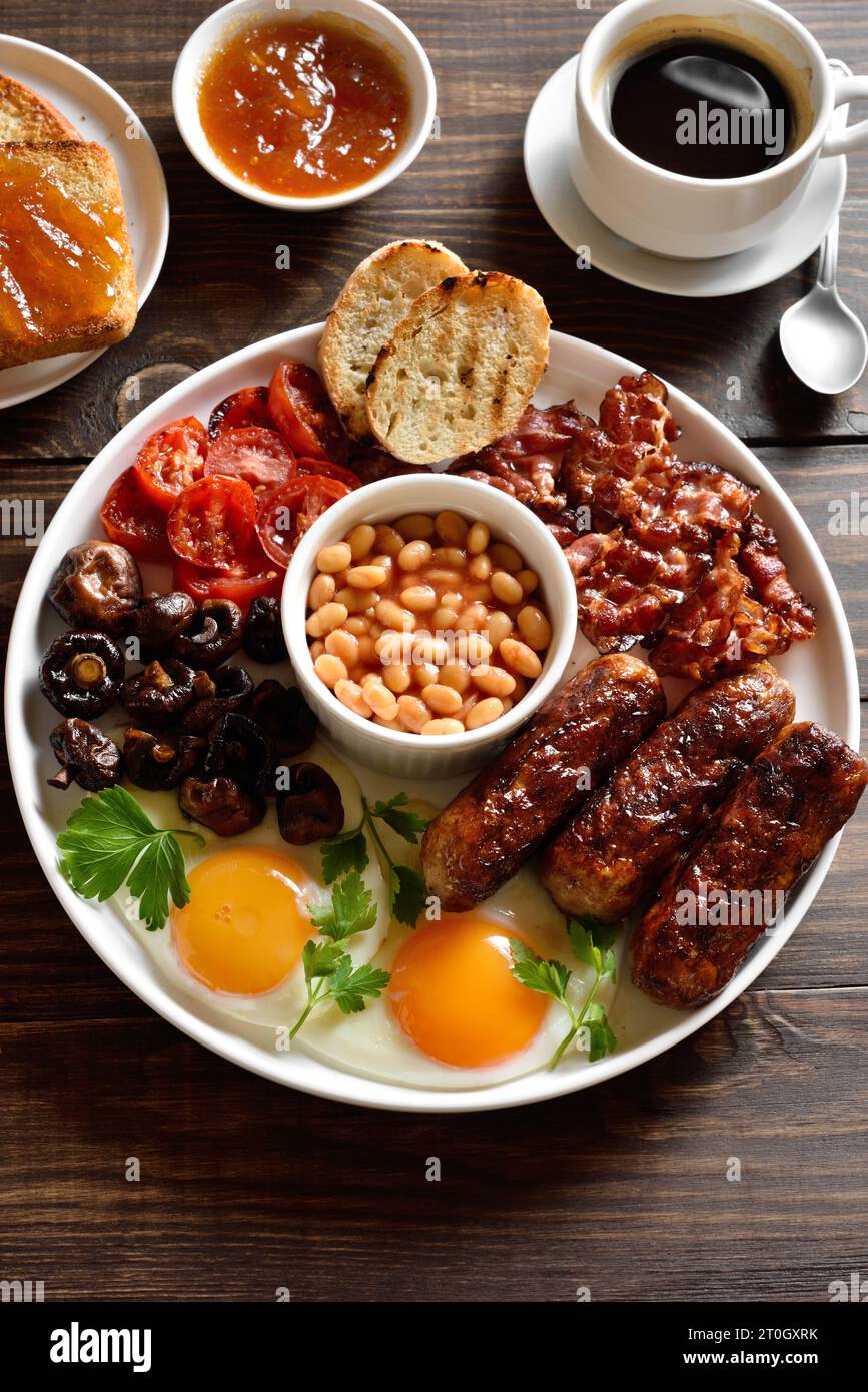 Colazione inglese completa con uova fritte, pancetta, salsiccia, fagioli e funghi su un tavolo di legno. Foto Stock