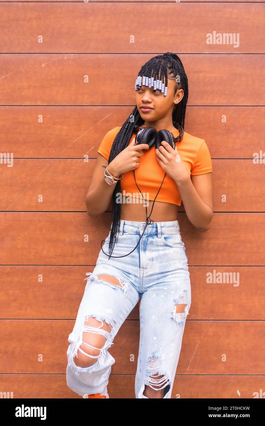 Stile di vita con una giovane ballerina in trappola in città. Ragazza nera del gruppo etnico africano con t-shirt arancione e pantaloni da cowboy, con cuffie su una Foto Stock