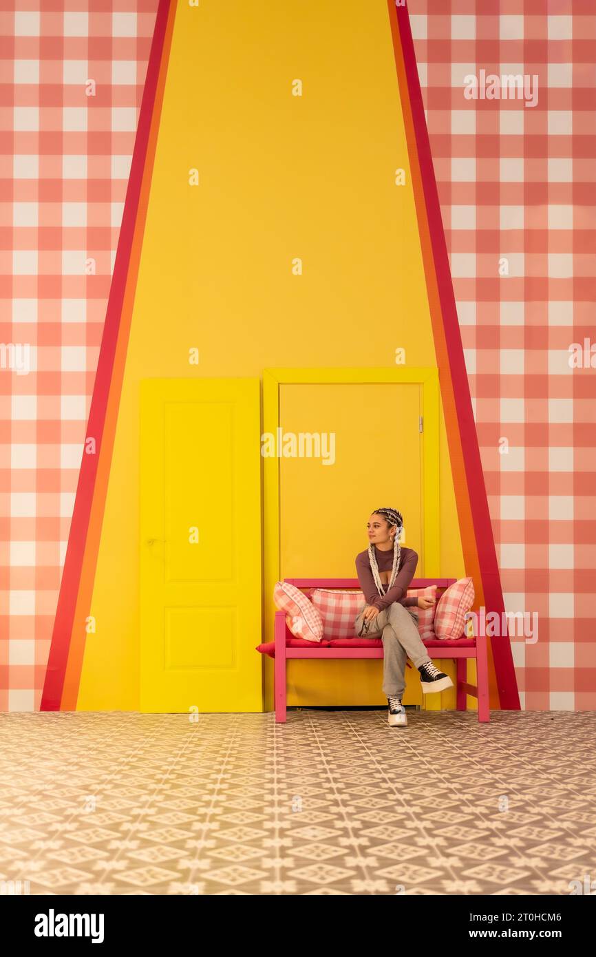 Giovane ragazza con trecce bianche su sfondo giallo, seduta su una panca rosa. Foto verticale, copia e incolla spazio Foto Stock