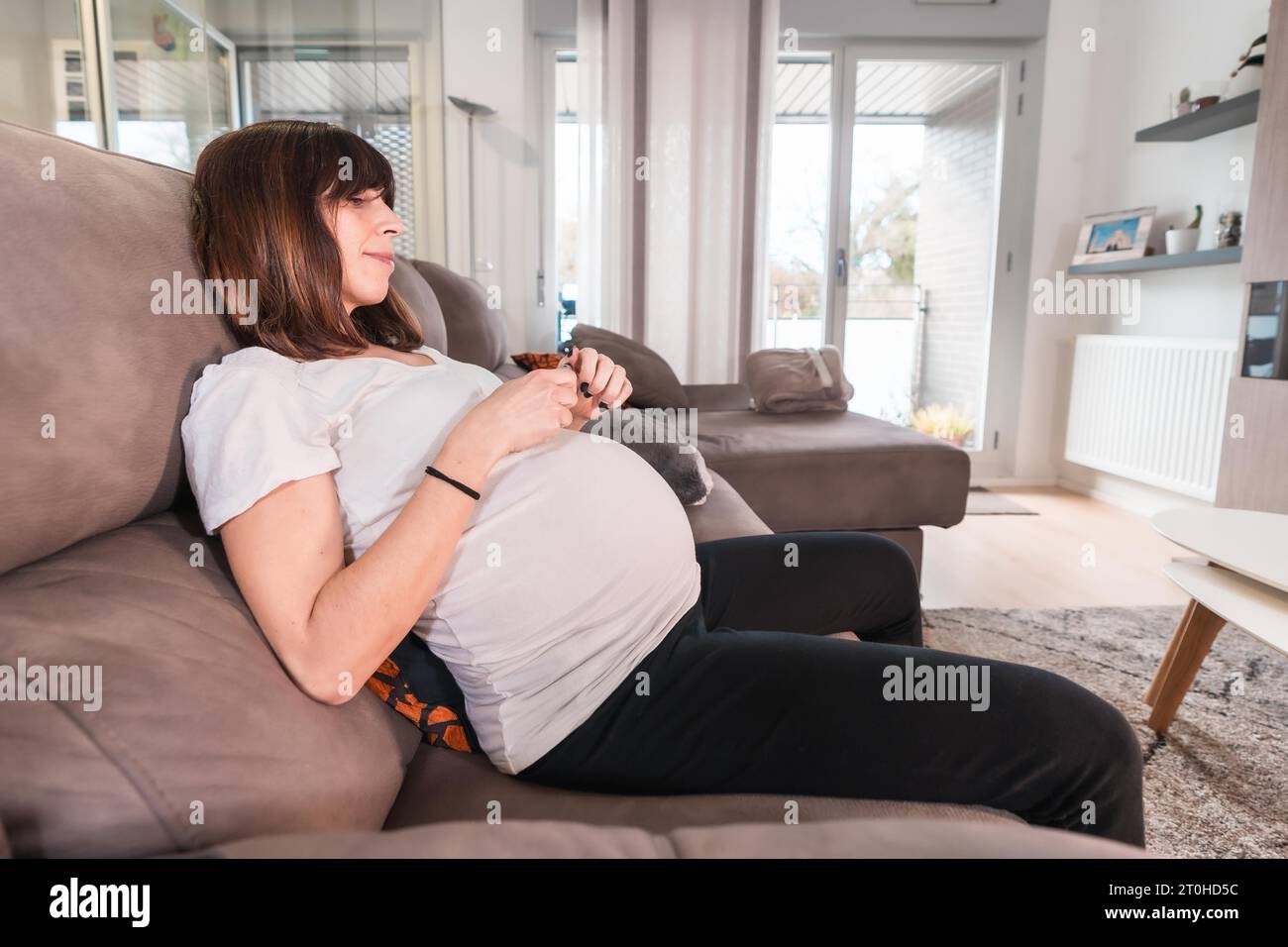 Giovane donna incinta con diabete gestazionale autotest per controllare lo zucchero sul divano a casa, misurando la goccia di sangue dal dito Foto Stock