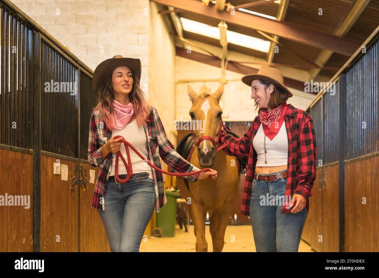 Due donne cowgirl che si divertono e ridono con un cavallo in una stalla, con abiti sudamericani Foto Stock