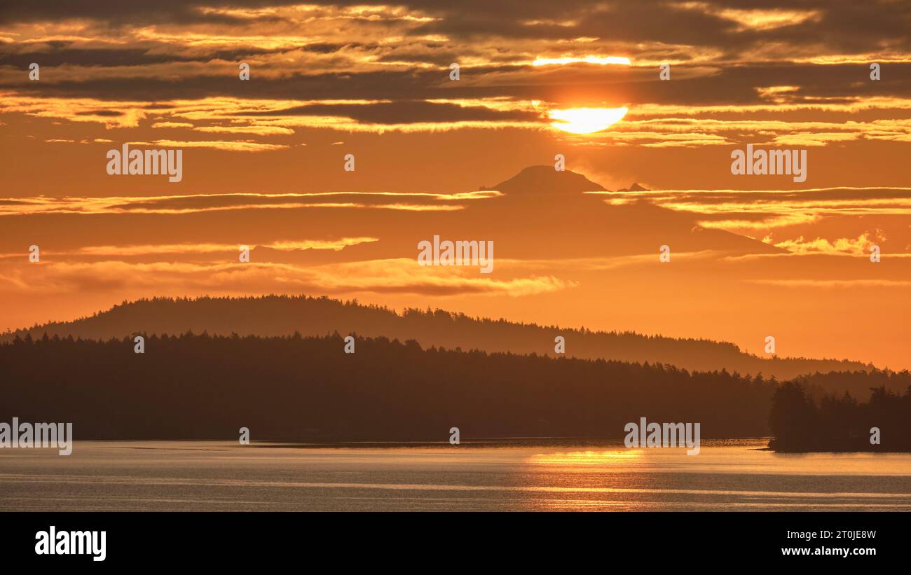 Il sole come una grande palla gialla sopra il monte Baker a 130 km di distanza è circondato da nuvole e da un cielo arancione come visto sull'acqua dall'Isola di Vancouver. Foto Stock