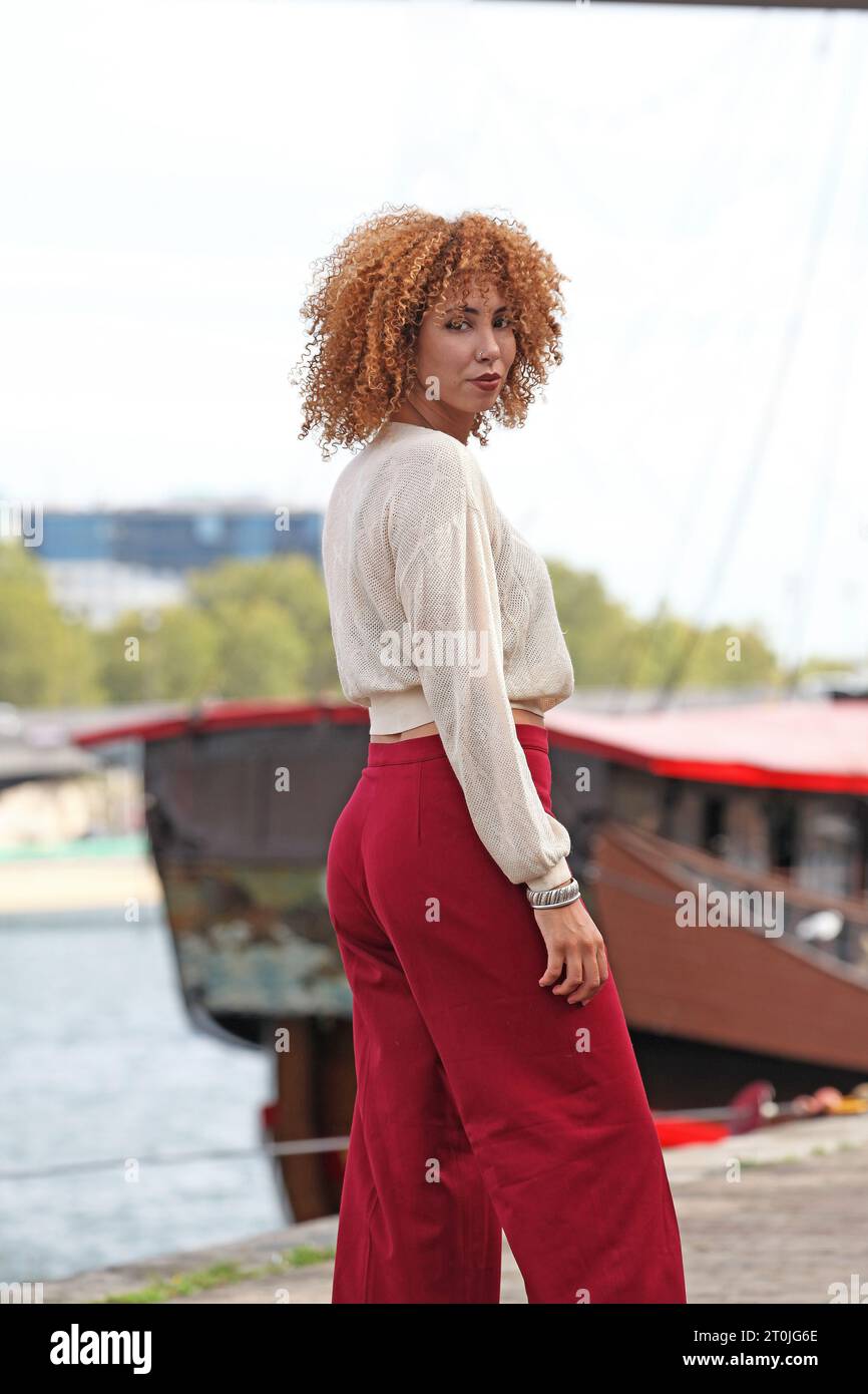 donna con capelli ricci e pantaloni rossi in posa su un molo vicino a una barca Foto Stock