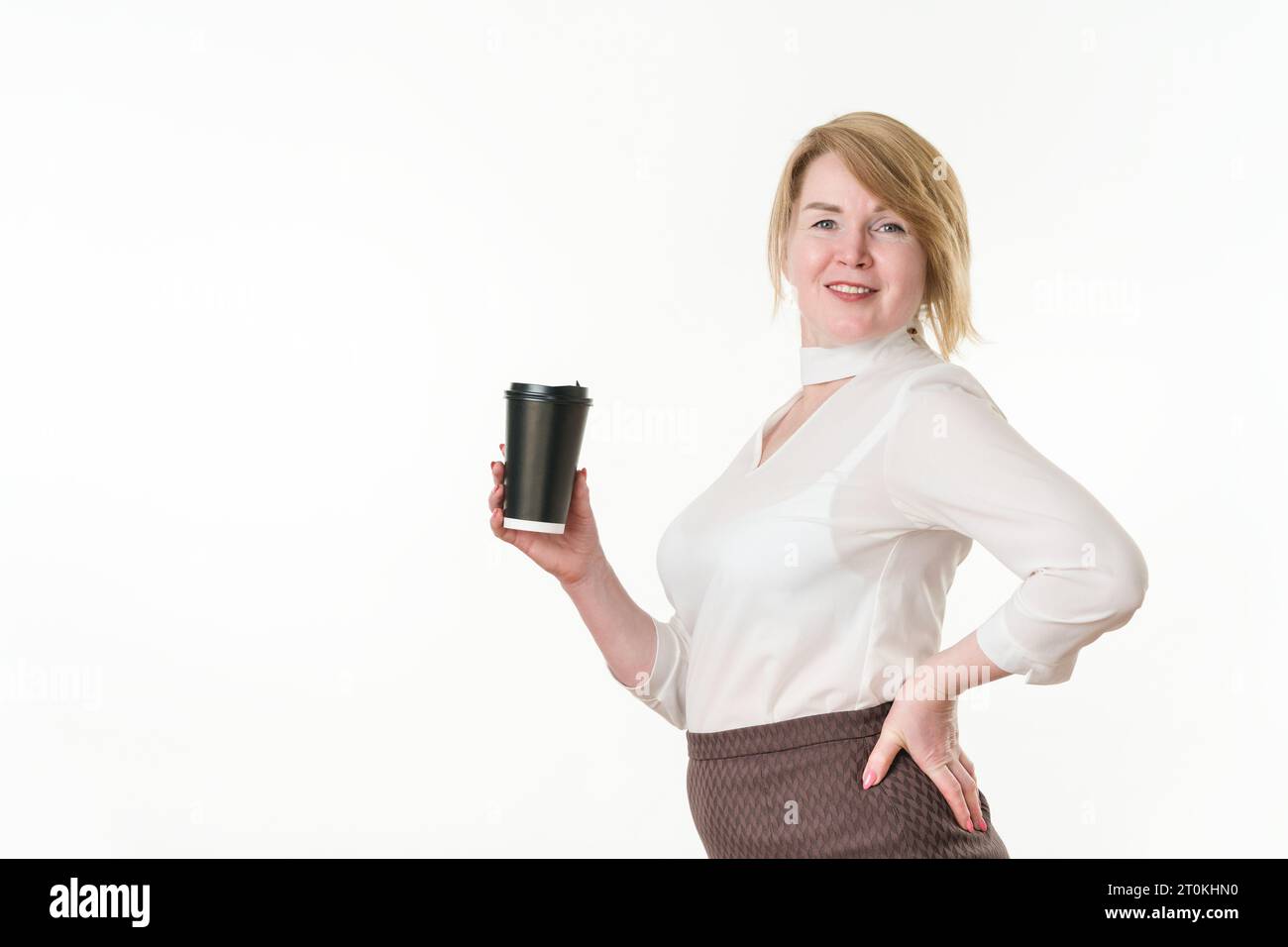 Donna bionda matura che tiene in mano una tazza di carta nera usa e getta per caffè o tè, l'altra mano è in vita. Donna che guarda la telecamera. Donna sorridente Foto Stock