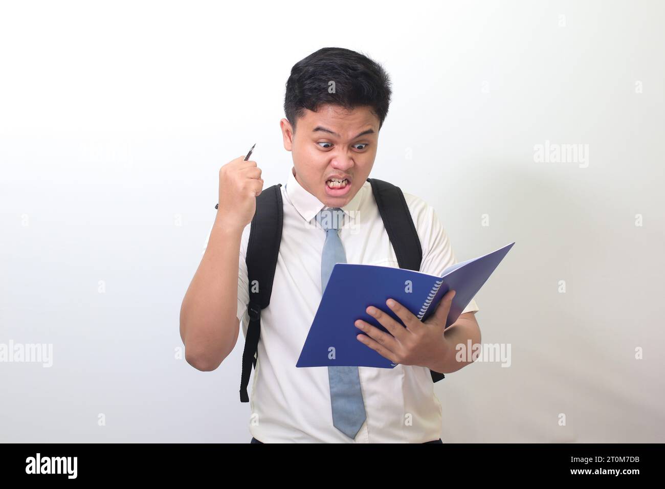 Studente delle superiori indonesiane che indossa un'uniforme bianca con cravatta grigia su un quaderno usando una penna con un'espressione infastidita e frustrata. Foto Stock