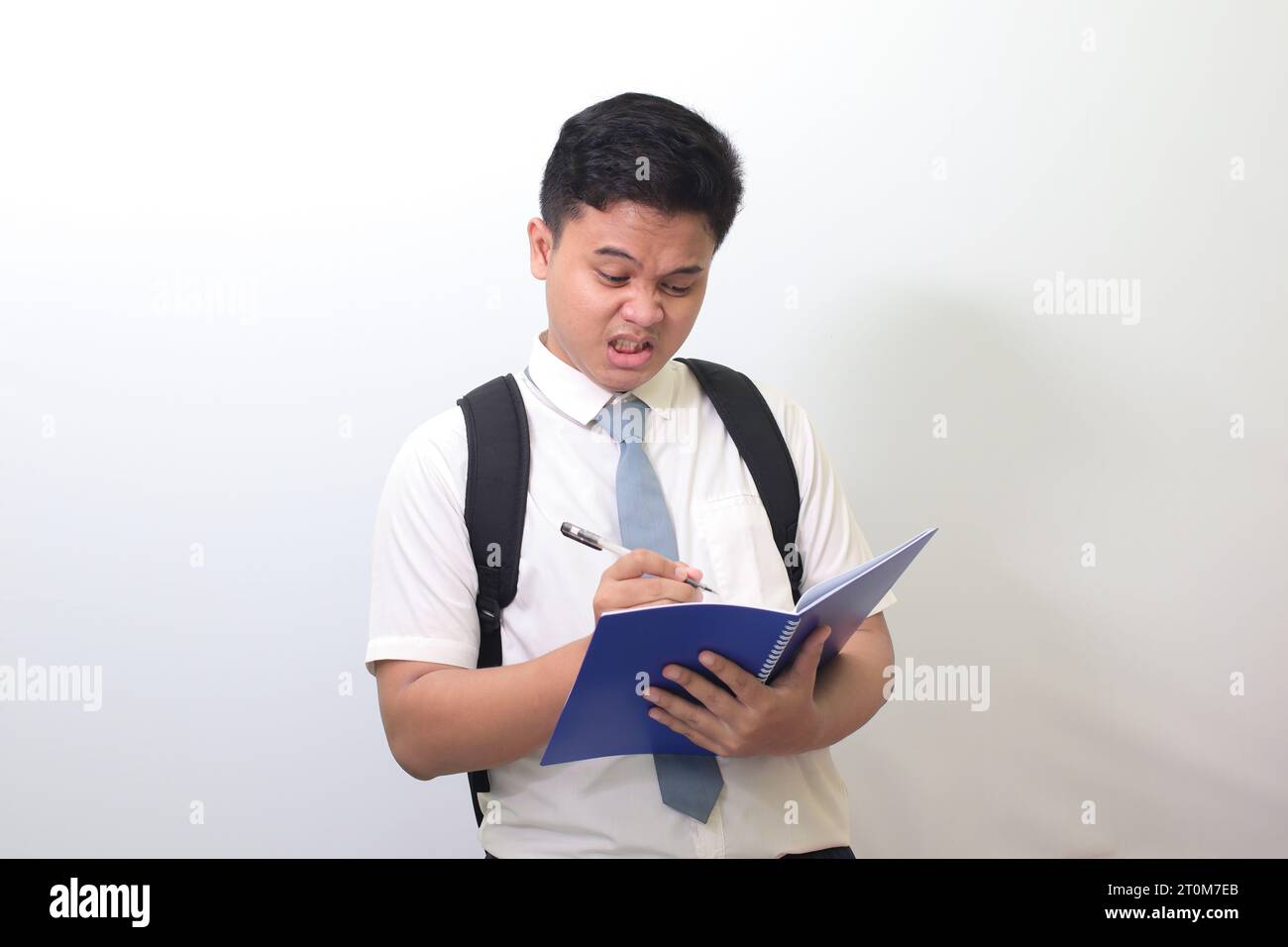 Studente delle superiori indonesiane che indossa un'uniforme bianca con cravatta grigia su un quaderno usando una penna con un'espressione infastidita e frustrata. Foto Stock