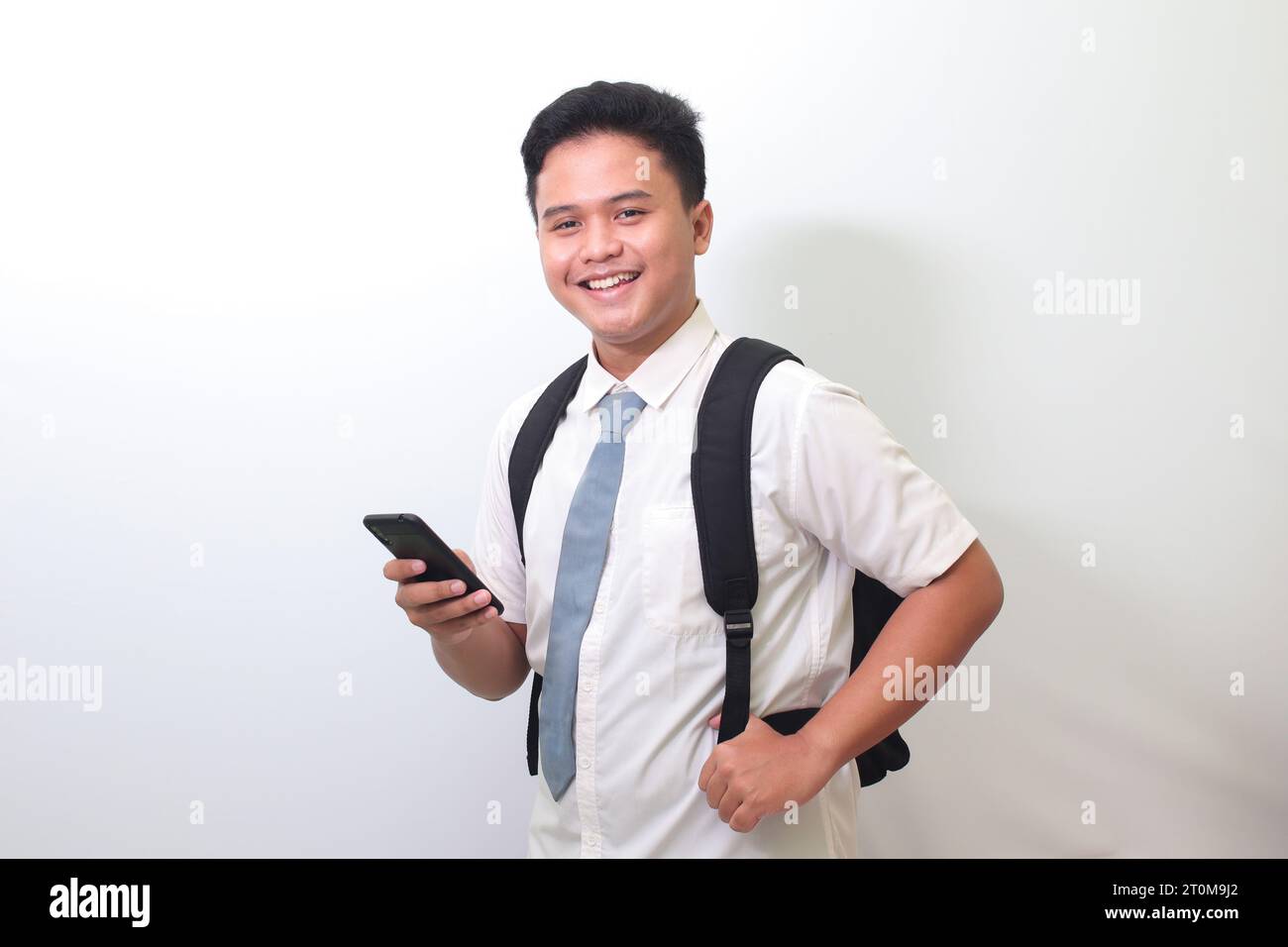 Studente indonesiano di scuola superiore che indossa una maglietta bianca in uniforme con cravatta grigia che mostra un'espressione scioccata del volto mentre tiene un telefono cellulare. Isolato Foto Stock