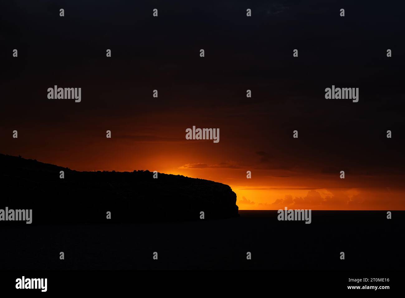 Il caldo bagliore di sole del mare si alza attraverso il buio del cielo, del mare e delle scogliere. Immagine minimalista a basso contrasto scattata sulla costa sud di mal Foto Stock