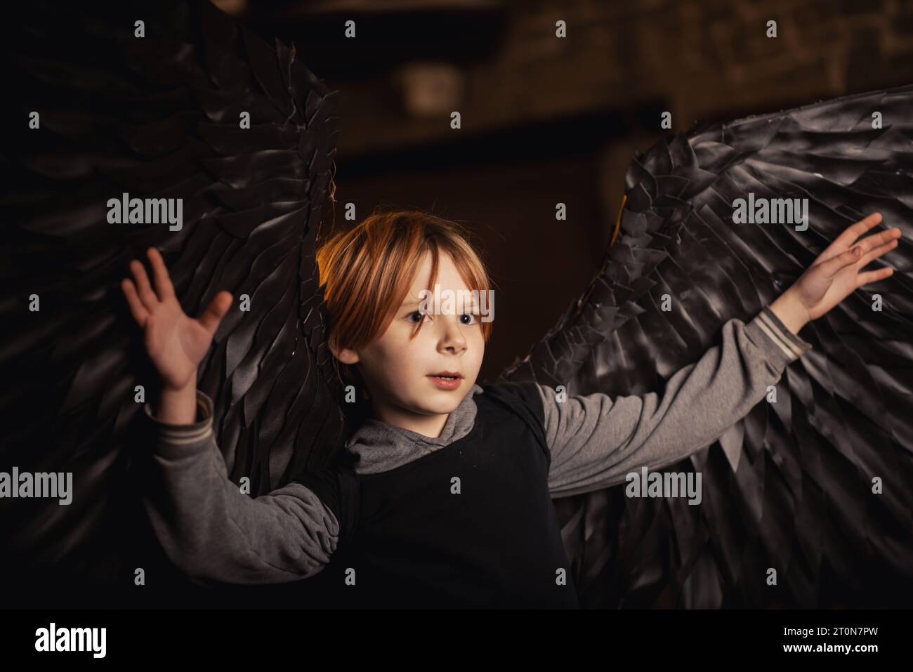 Un ragazzo con ali d'angelo nero siede in una stanza buia. Le sue ali sono l'unica cosa che lo fa risaltare nell'oscurità. Foto Stock