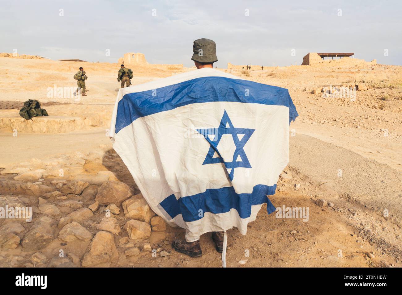 Vista posteriore del soldato israeliano con bandiera bianca e blu di Israele sullo sfondo del territorio fortificato di Masada. Uomini militari che proteggono profanati Foto Stock