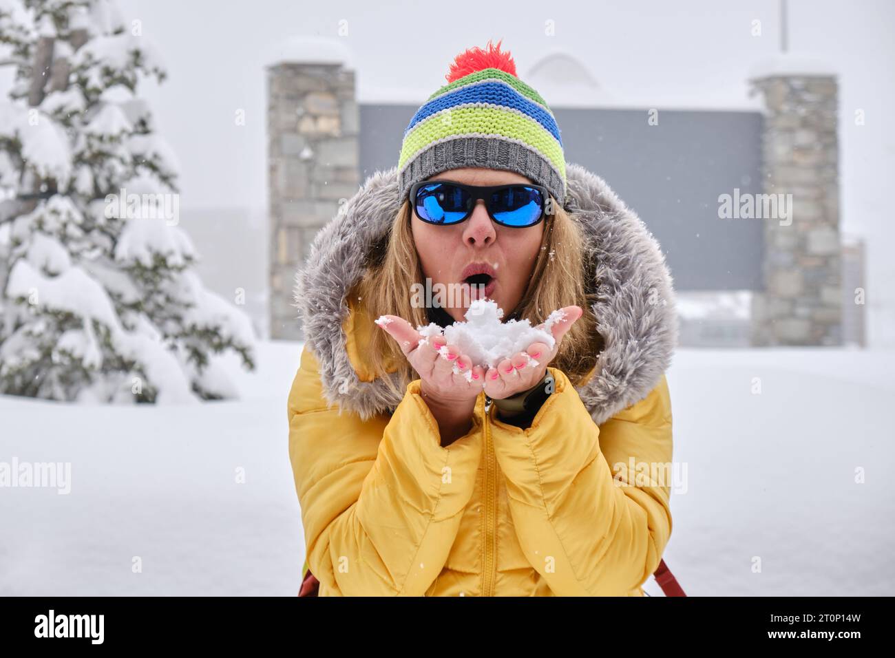Donna che soffia neve dalle mani, indossa abiti colorati di colore giallo in inverno. Felicità, gioia, giocoso. Foto Stock
