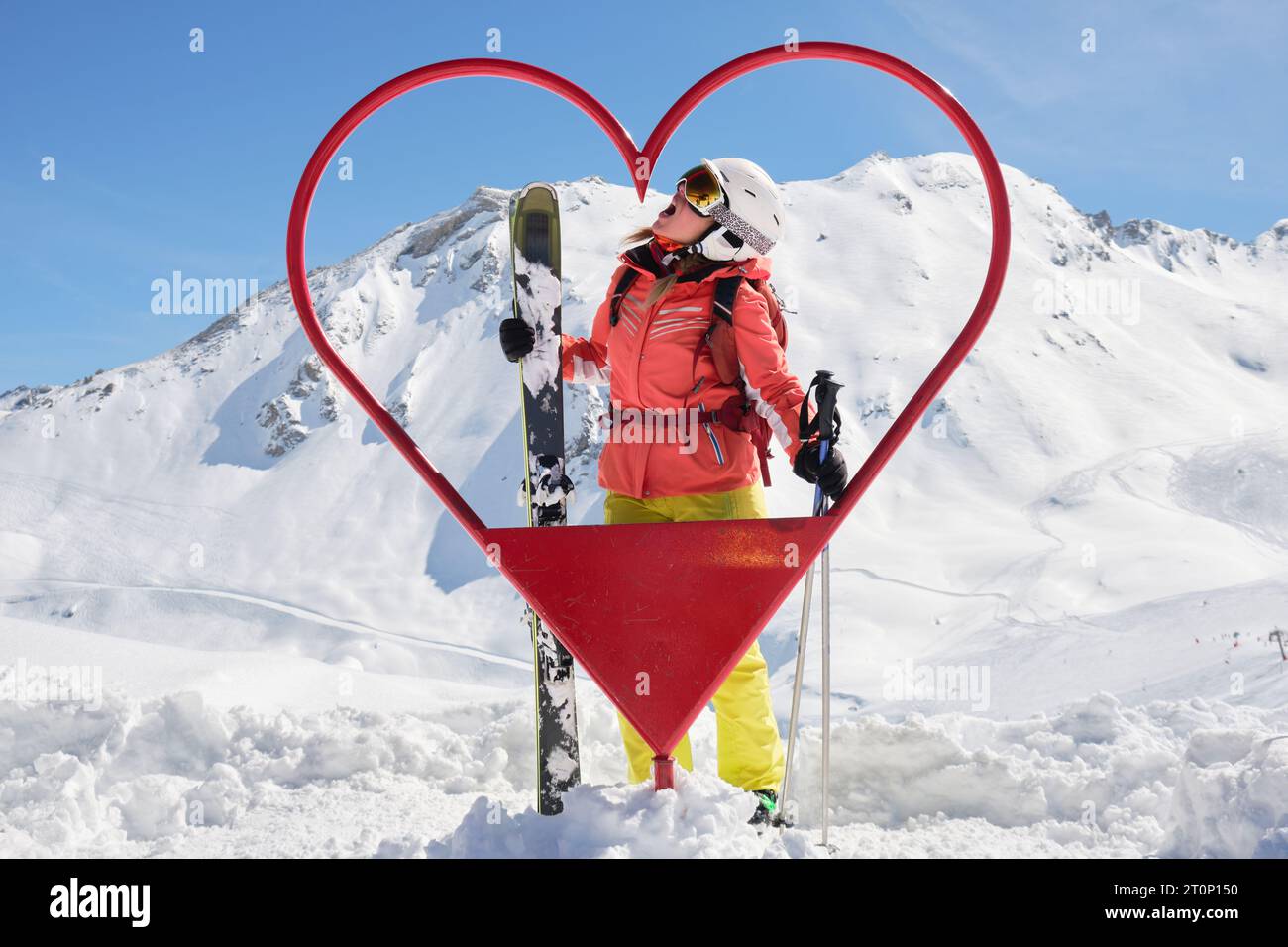 Sciatore donna posa all'interno di una cornice a forma di cuore a Tignes - Val d'Isere località sciistica, in Francia, con tutti gli sci montani e vestito rosso, in una giornata di sole. Touris Foto Stock