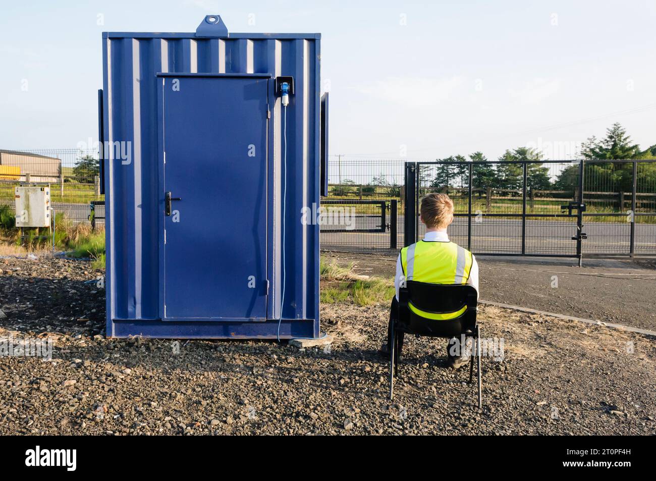 Una guardia di sicurezza si siede su una sedia accanto alla sua capanna all'interno di un recintato composto Foto Stock