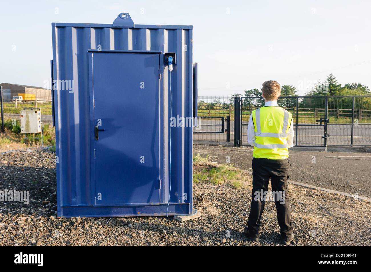 Una guardia di sicurezza si erge accanto alla sua capanna all'interno di un recintato sito in costruzione composto Foto Stock