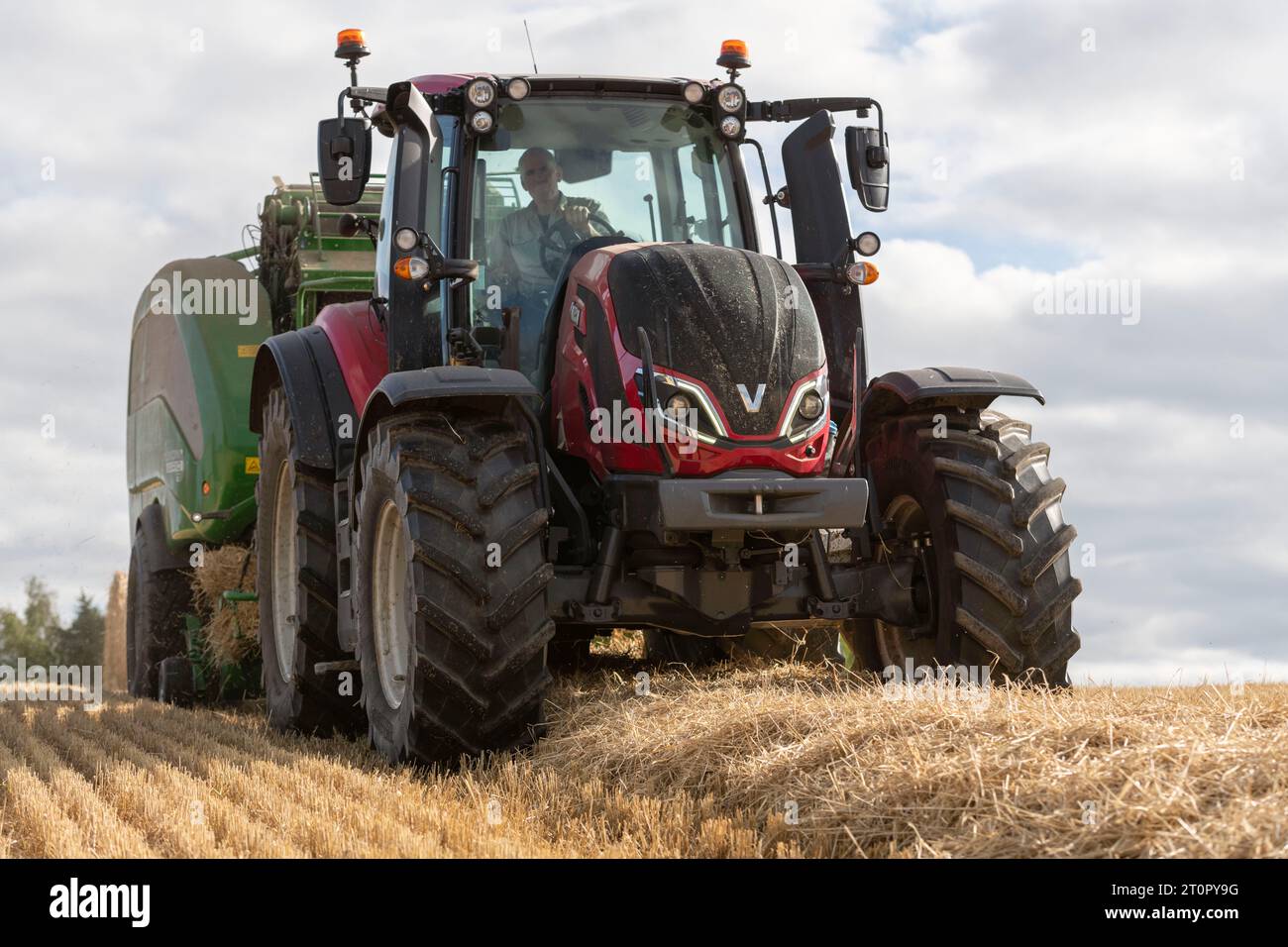 Un trattore Valtra t195 rosso che traina un'imballatrice in un campo di paglia d'orzo visto dalla parte anteriore e da un punto di vista basso Foto Stock