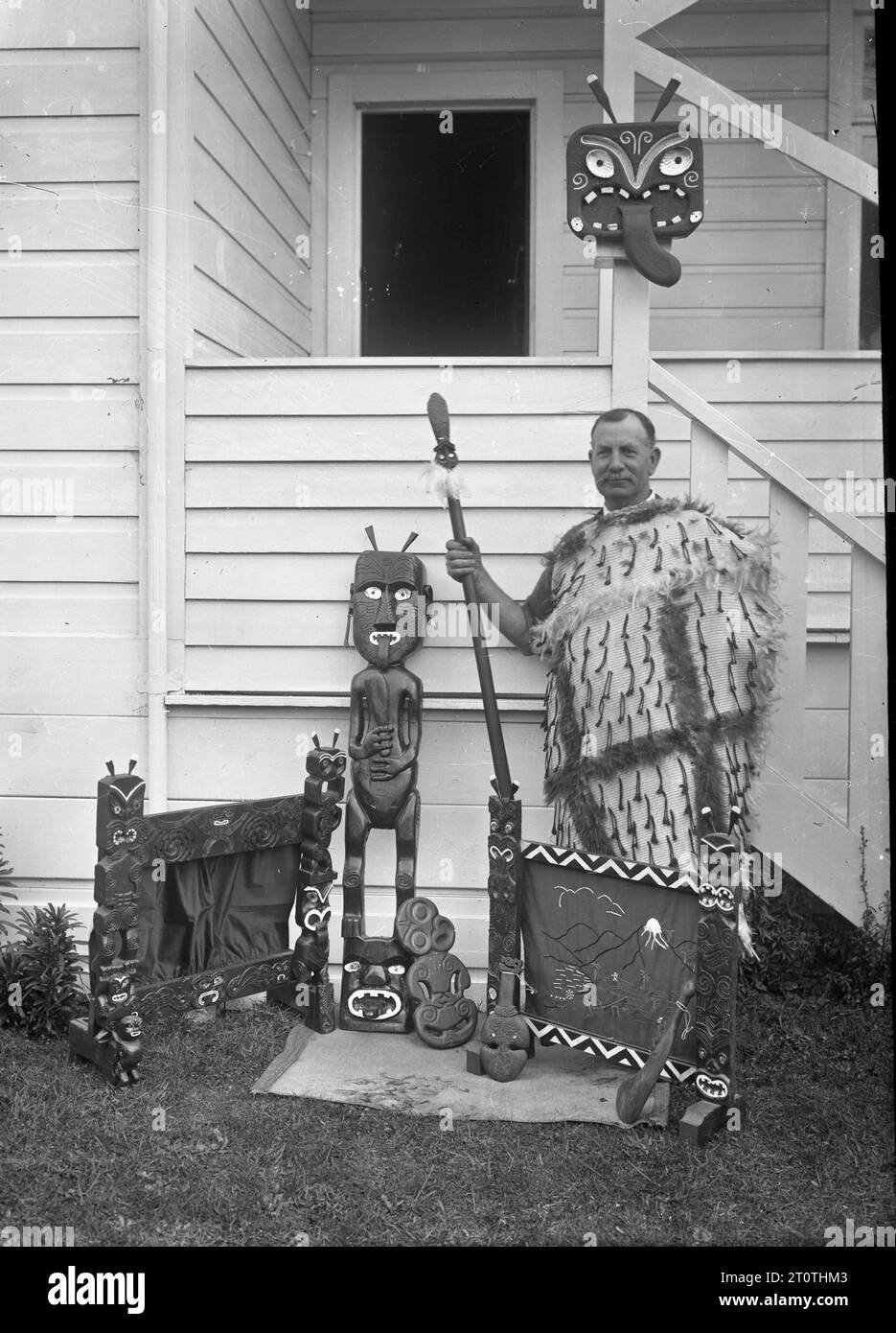 Albert Percy Godber (fotografo neozelandese) - indossa un mantello Maori, con in mano una taiaha, accanto a una collezione di sculture Maori - 1932 Foto Stock