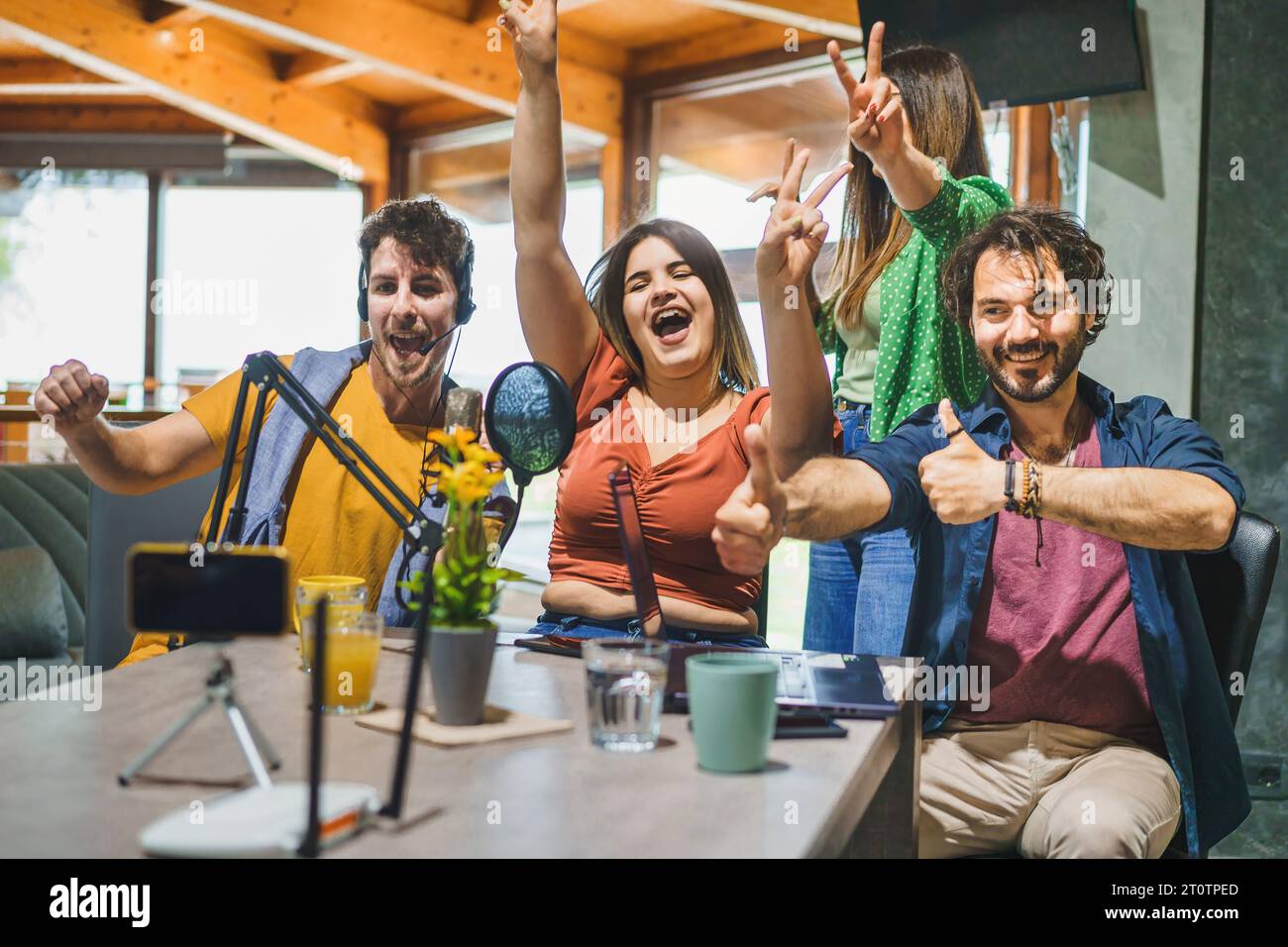 Un gruppo di quattro creatori di contenuti festeggia e si muove con gioia verso uno smartphone montato su un treppiede, con un microfono davanti, come Foto Stock