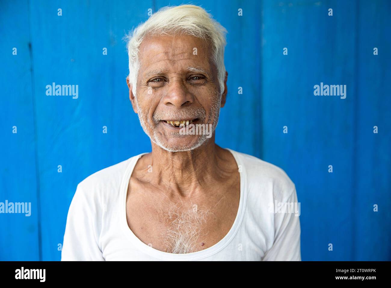 Un uomo del posto sorridente davanti a una parete di legno di colore blu nella sua casa in un piccolo villaggio sull'isola di Majuli, il fiume Brahmaputra, Assam, India Foto Stock
