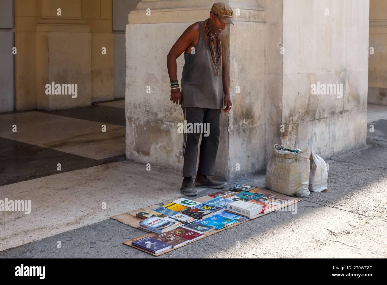 Un uomo cubano vende media scritti sul marciapiede. I muri intorno a lui sono sporchi e rovinati. Foto Stock