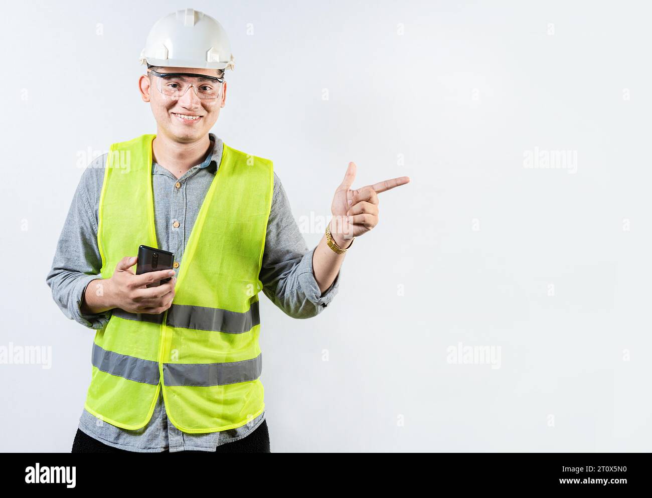 Ingegnere sorridente che tiene il cellulare e indica un annuncio. Giovane ingegnere latino che tiene il telefono e punta verso uno spazio vuoto Foto Stock
