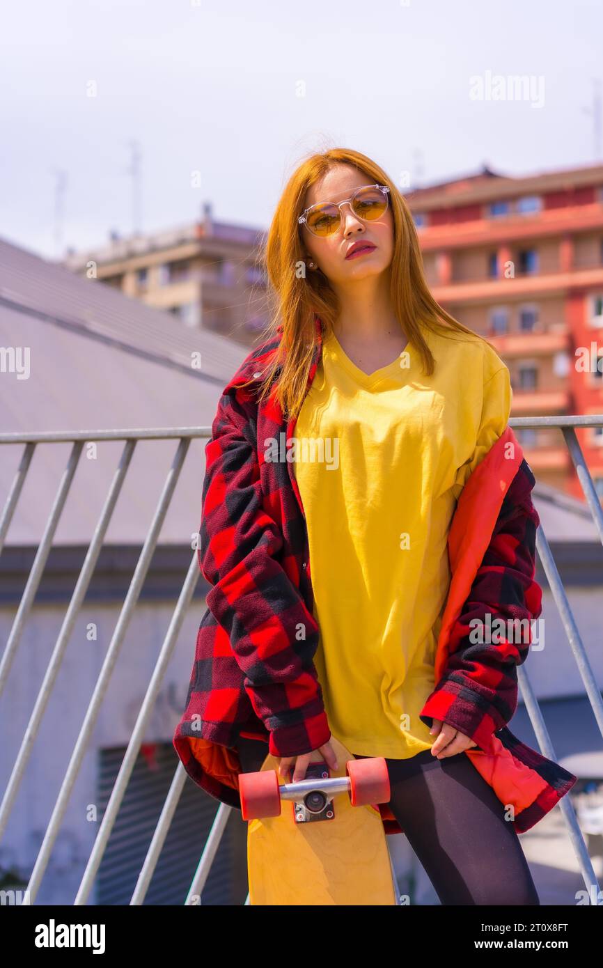 Donna skateboarder con una t-shirt gialla, una camicia a quadri rossa e occhiali da sole, in posa con skateboard, foto verticale Foto Stock