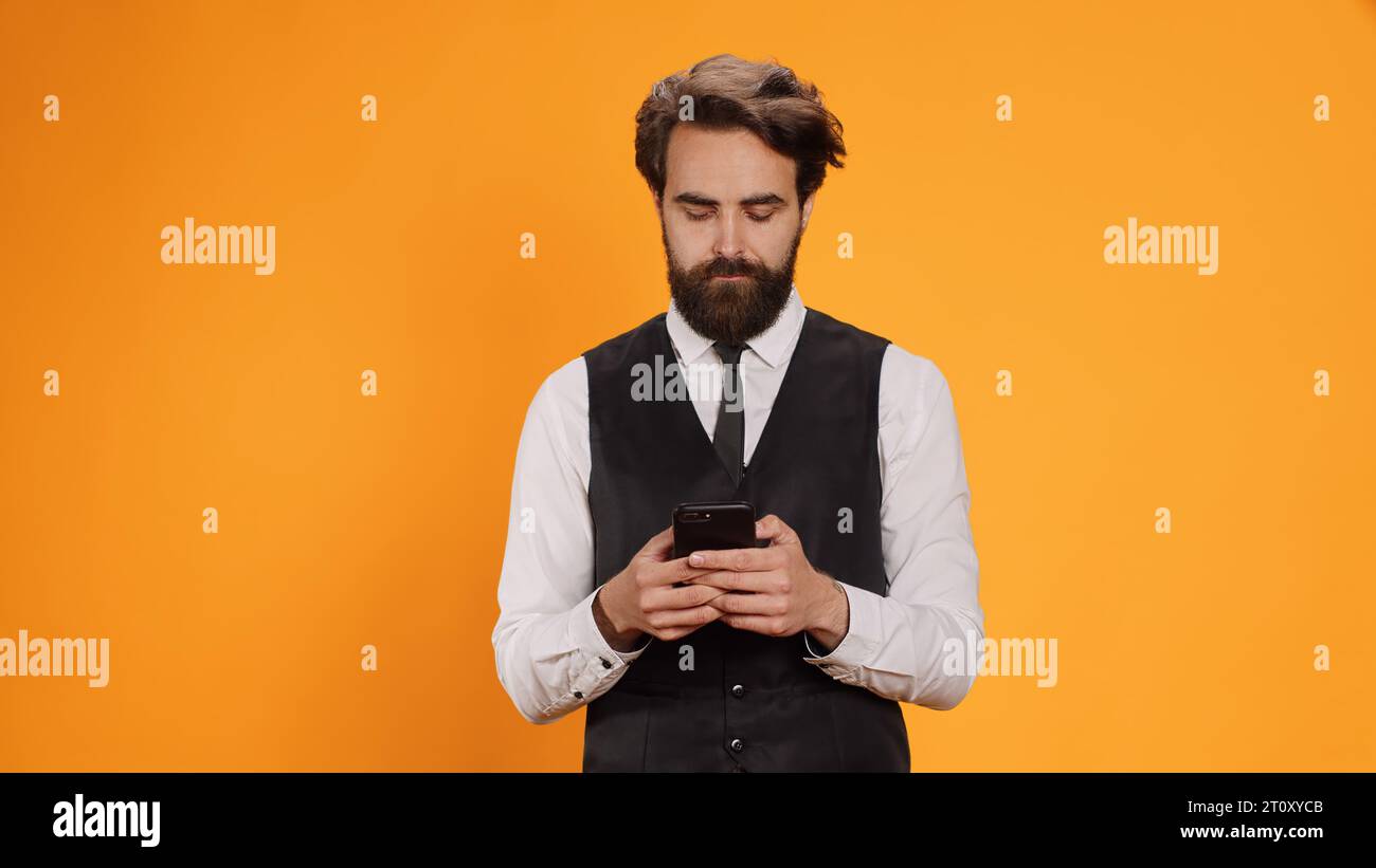 Un cameriere competente controlla lo smartphone, pronto a servire i clienti al tavolo. Il personale sofisticato e alla moda che lavora nel settore culinario dipende dall'applicazione per telefoni cellulari e dalla messaggistica. Foto Stock