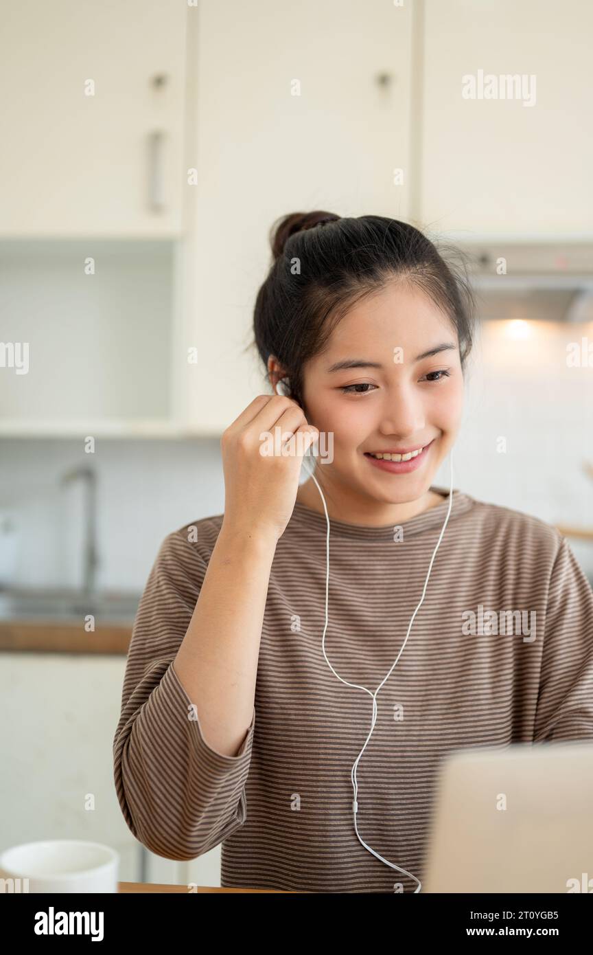 Un'attraente giovane donna asiatica lavora da casa, indossa gli auricolari e usa il suo laptop a un tavolo in cucina, organizza una riunione online, o s Foto Stock