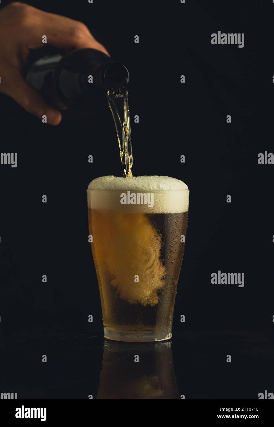 Shot frontale di un bicchiere di birra e di una mano che versa la birra su uno sfondo nero con una schiuma che forma il ruscello. Foto Stock