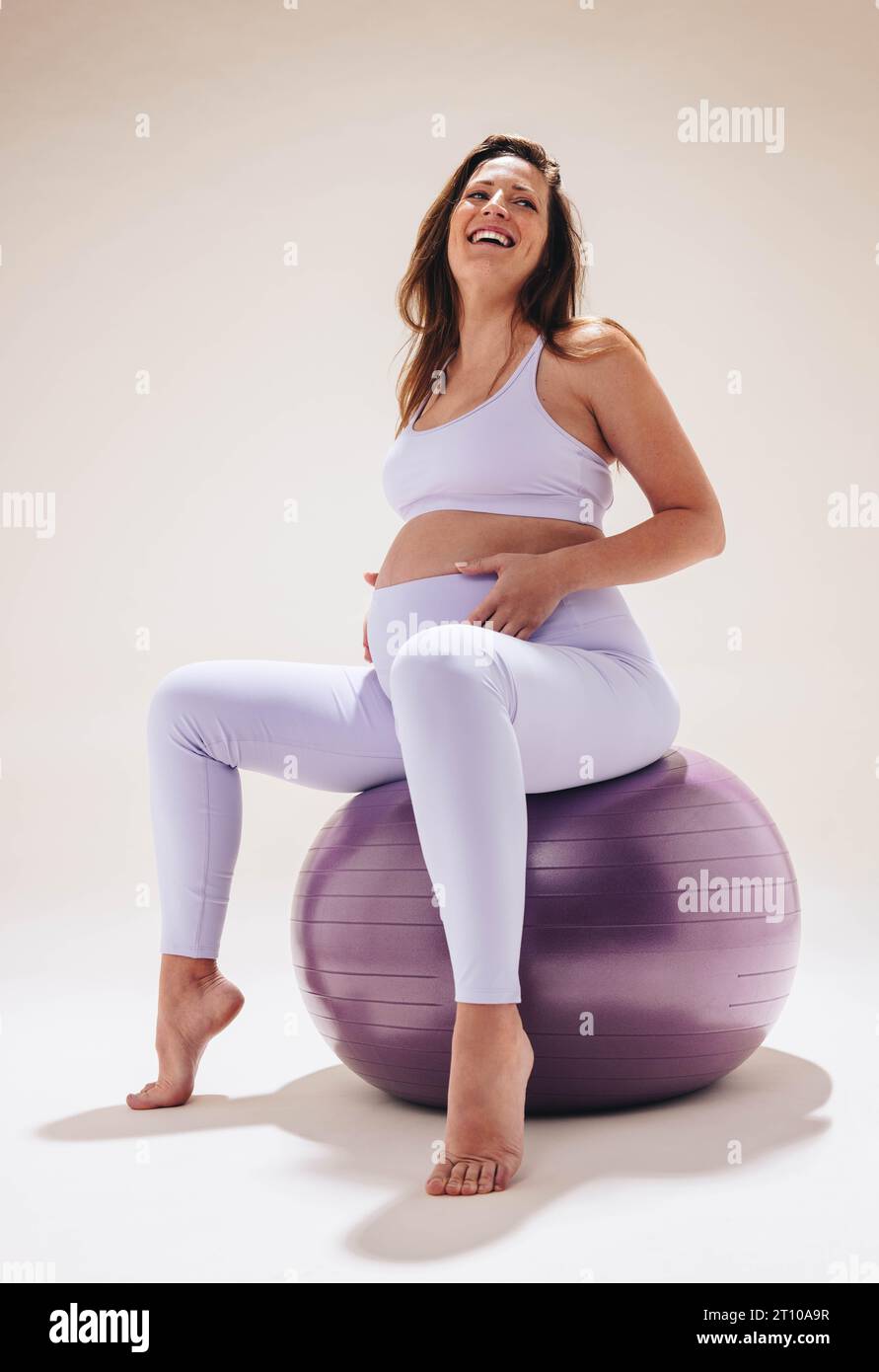 Una donna incinta felice nel suo terzo trimestre pratica yoga prenatale su una palla di stabilità in studio. Sorride e si prende cura del suo corpo, abbracciando Foto Stock